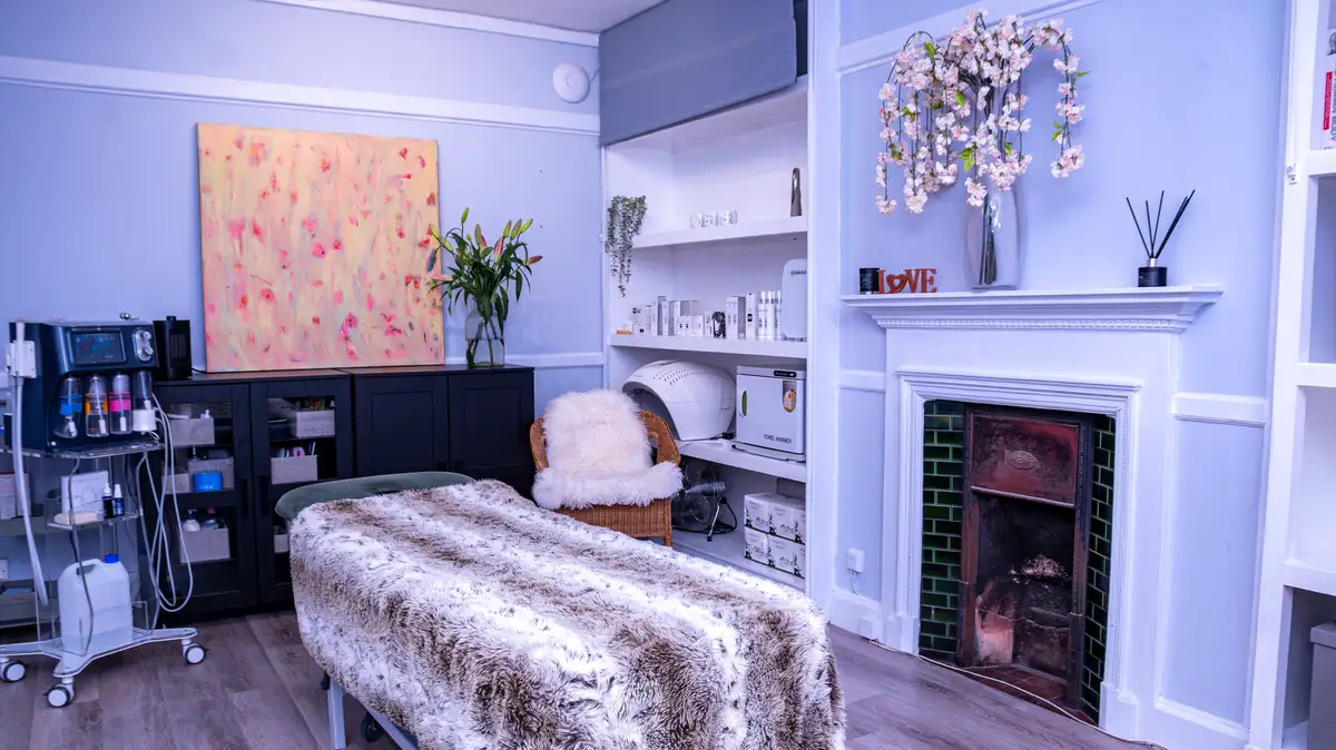 A treatment room with a massage table covered in a fur blanket, a fireplace, shelves with decor, flowers, and spa equipment.