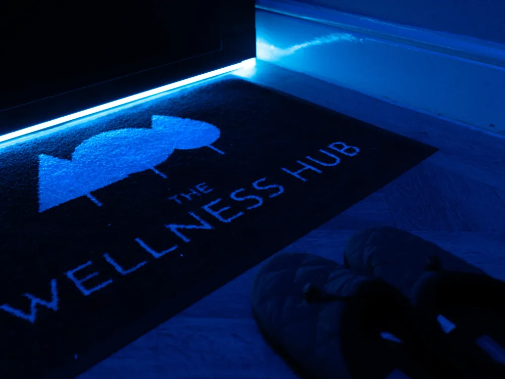 A mat reading The Wellness Hub is placed by the opening of the full body cryotherapy chamber. A pair of slippers sits on the floor nearby.