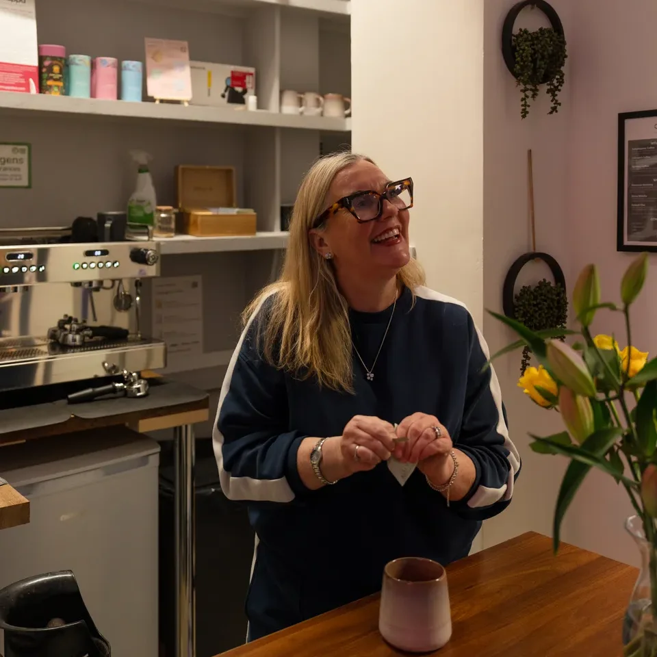 Emma stands smiling behind a wooden counter in a coffee shop, with a cup in front of her and shelves with coffee supplies in the background.