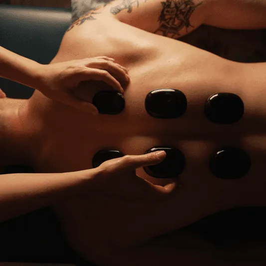 Person receiving a hot stone massage on their back, with several smooth black stones placed in two rows, and a tattoo visible on their upper arm.