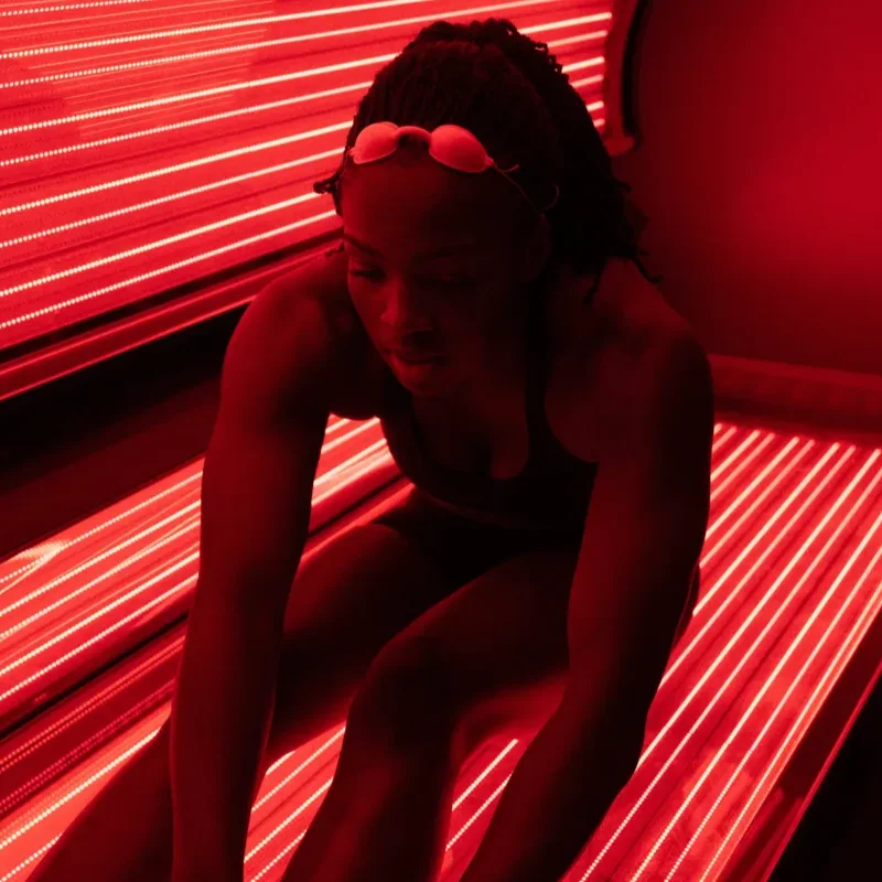 A person in swimwear and goggles sits inside a sunbed illuminated by red lights.