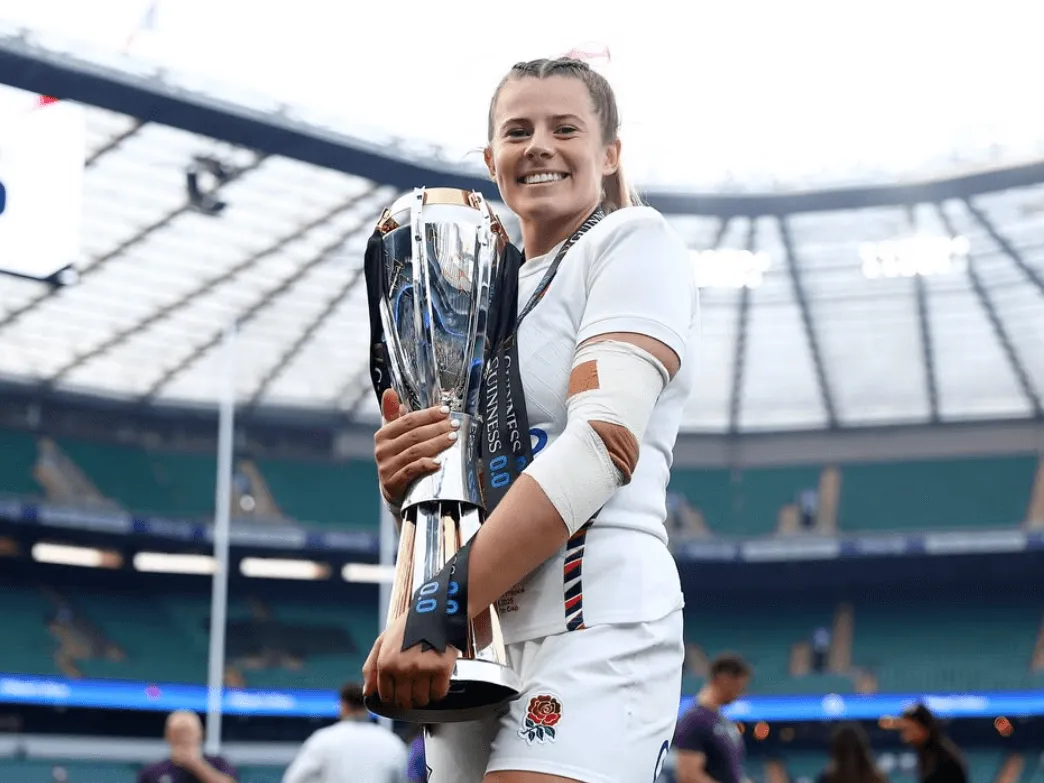Zoe Harrison in an England kit stands in a stadium, smiling and holding a large trophy.