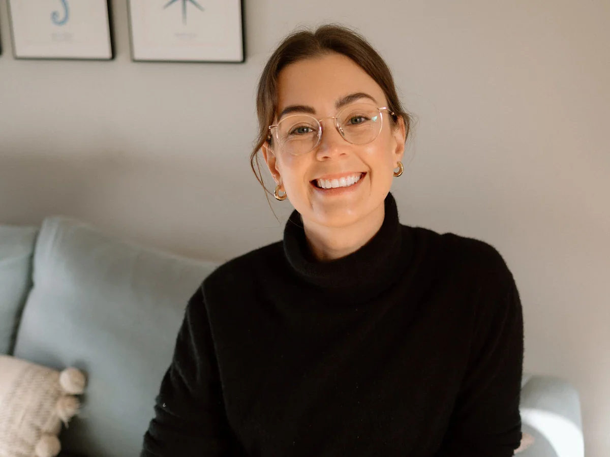 Dr Kate Watchorn, wearing glasses, wearing a black polo neck jumper, smiles whilst sitting on a light-coloured sofa in a softly lit room.