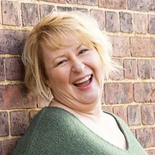 Woman with short blonde hair smiling and leaning against a brick wall, wearing a green jumper.