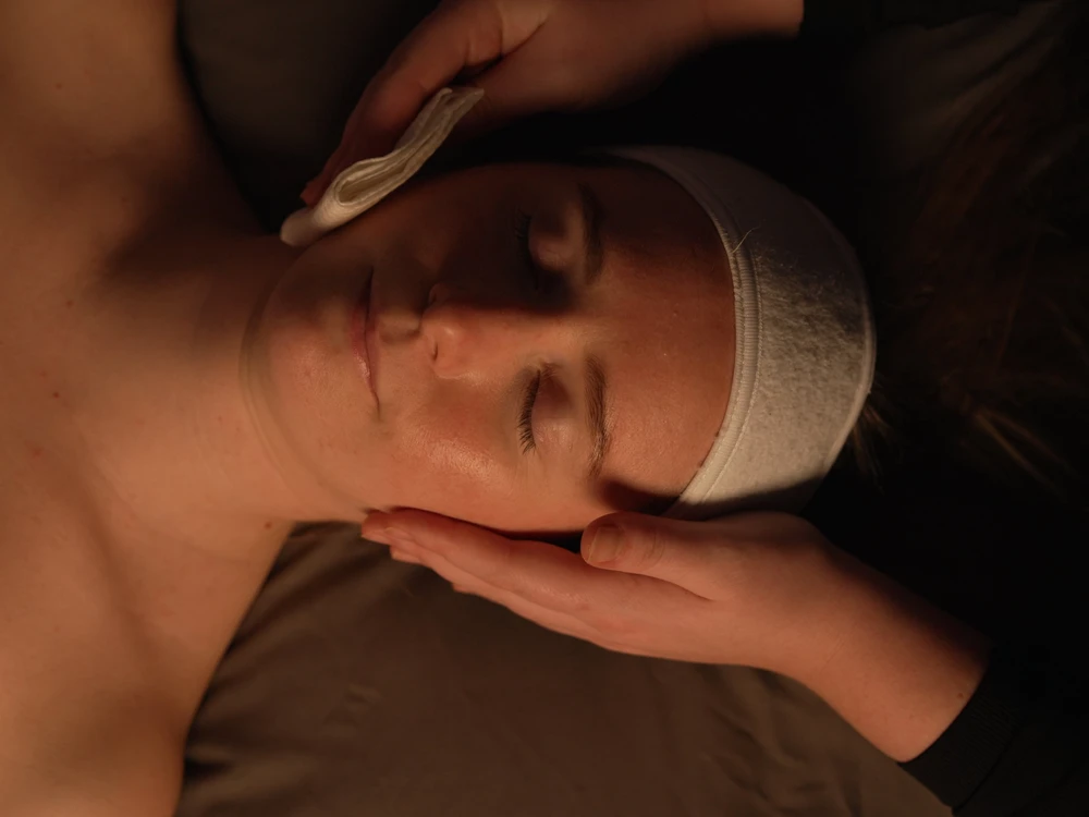A person lies with eyes closed while another person gently cleanses their face with a cotton wool pad, suggesting a facial treatment or spa session.