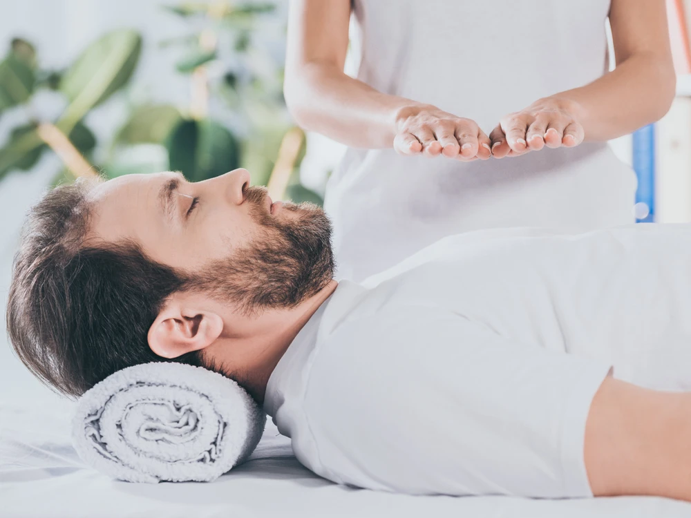 A man lies on a table with a towel under his head whilst another person holds their hands above his chest, performing an energy healing session.