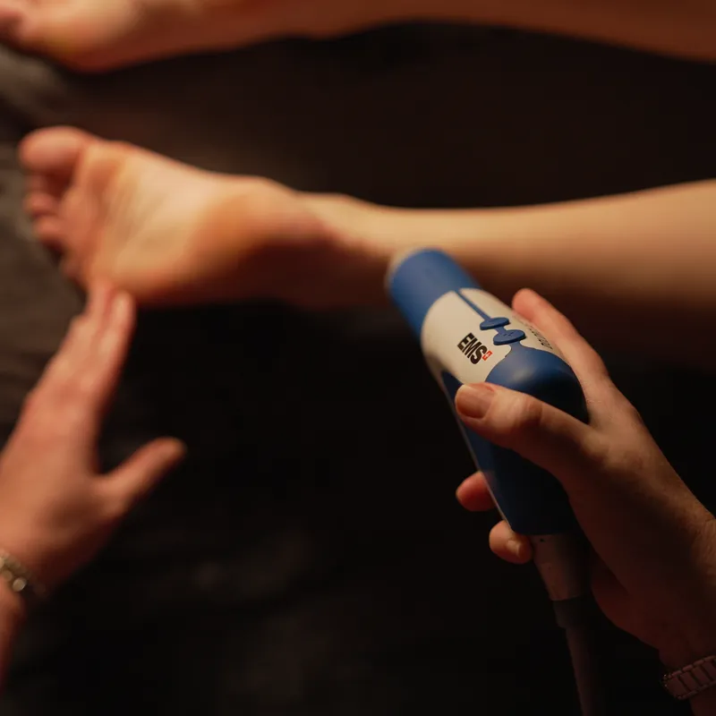 A person uses a shockwave therapy device on the sole of another person's foot, while the other hand supports the foot.