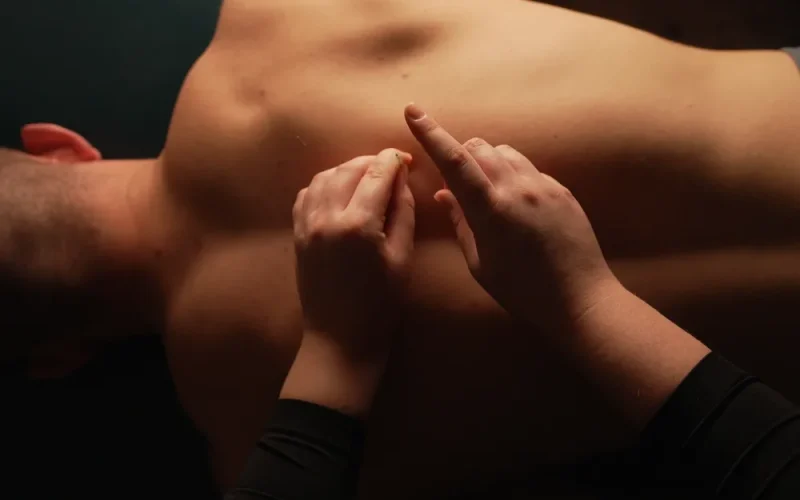 A person receives a back massage while lying face down on a table; the masseur's hands are pressing into their upper back near a tattoo.