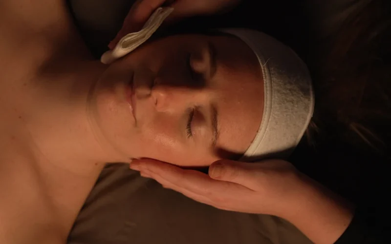 A person lies with eyes closed while another person gently cleanses their face with a cotton wool pad, suggesting a facial treatment or spa session.