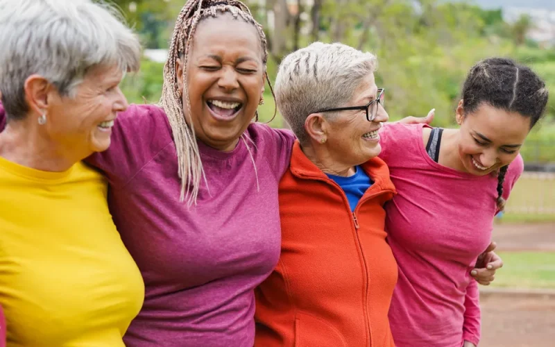 Four women stand outdoors, arms round each other, smiling and laughing together.