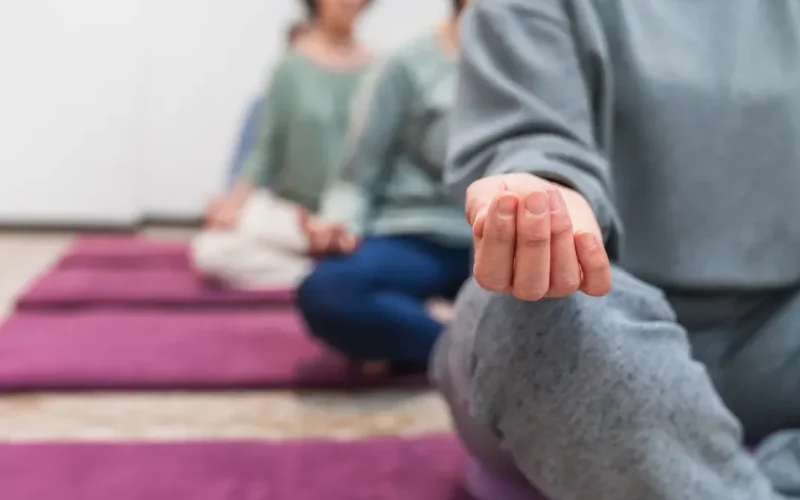 A group of people sit cross-legged on purple mats indoors, meditating with hands resting on knees. The focus is on the hand of the person in the foreground.