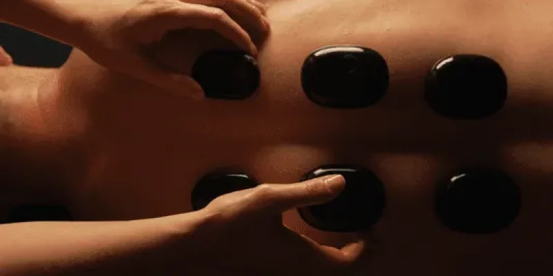 Person receiving a hot stone massage on their back, with several smooth black stones placed in two rows, and a tattoo visible on their upper arm.
