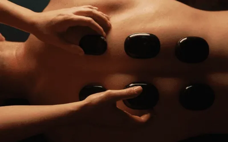 Person receiving a hot stone massage on their back, with several smooth black stones placed in two rows, and a tattoo visible on their upper arm.