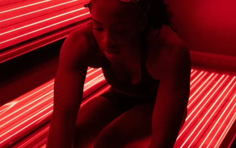 A person in swimwear and goggles sits inside a sunbed illuminated by red lights.