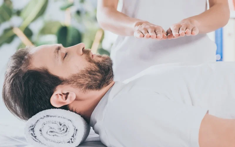 A man lies on a table with a towel under his head whilst another person holds their hands above his chest, performing an energy healing session.