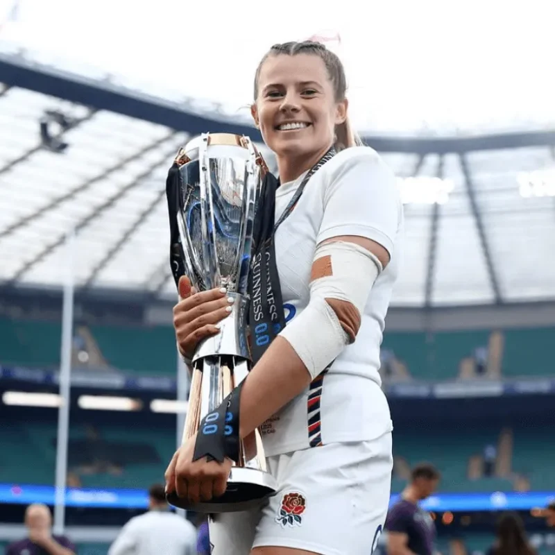 Zoe Harrison in an England kit stands in a stadium, smiling and holding a large trophy.