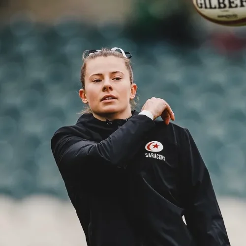 Zoe Harrison in a Saracens jacket prepares to catch a rugby ball during training, with stadium seats blurred in the background.